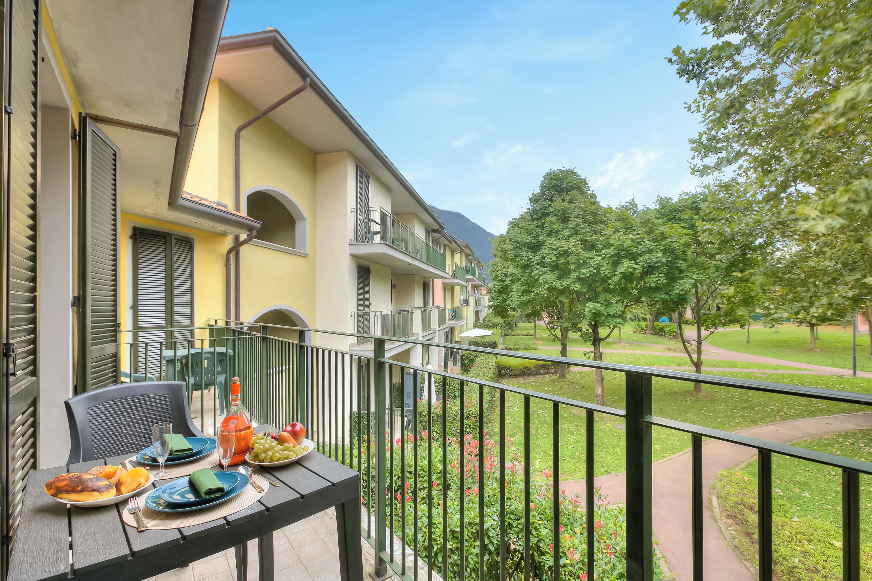 A balcony with a table set for breakfast, overlooking a garden and a row of buildings.
