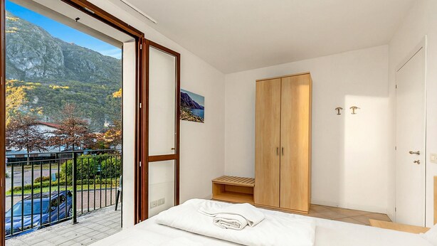 A bedroom with a bed, a wooden wardrobe, a small wooden stool, and a balcony with a view of mountains and a car.