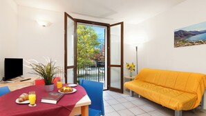 A room with a dining table set for breakfast, a sofa, a TV, and a view of trees and mountains through a glass door.
