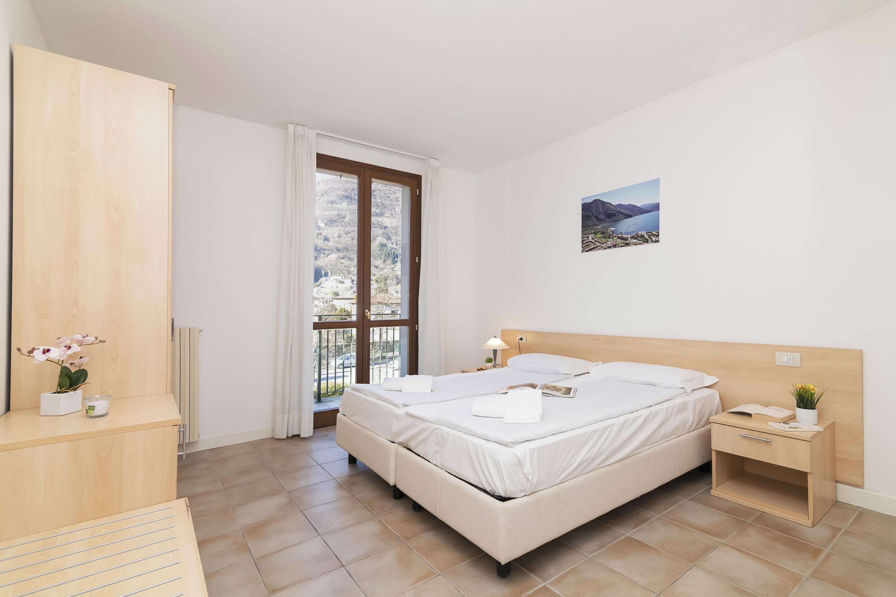 A bedroom with a large bed, a bedside table, a nightstand, a window with curtains, a radiator, and a wall-mounted picture.