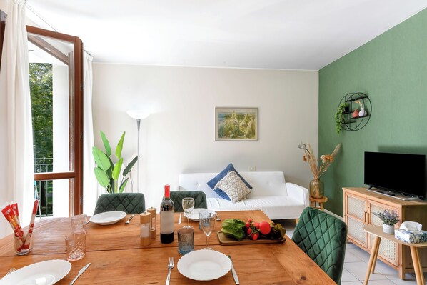 Comedor con una mesa de madera dispuesta para cuatro, un sofá blanco, un televisor y una pared verde con un cuadro.