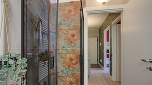 Un couloir avec une porte en verre donnant sur une salle de bain, une plante et une porte recouverte d’un papier peint à motifs floraux colorés.
