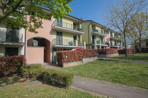 A row of residential buildings with balconies, a paved walkway, and well-maintained hedges.