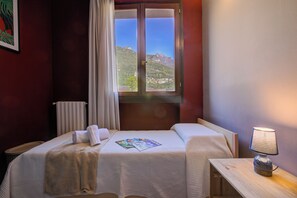 A hotel room with a bed, a bedside table with a lamp, a window with a view of mountains, and a radiator.
