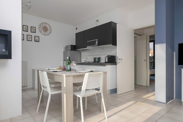 A modern kitchen with a dining table and chairs, a clock on the wall, and a view into the adjacent room.