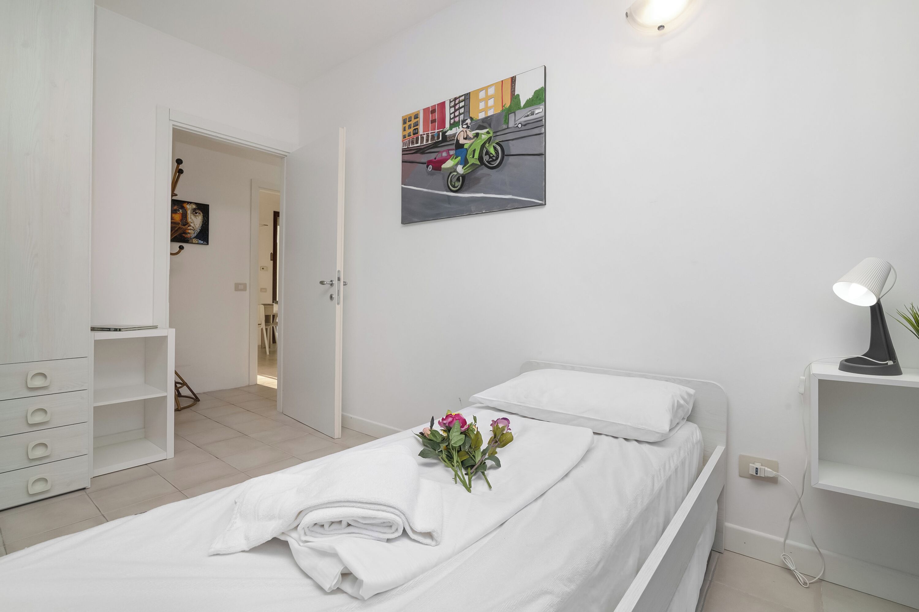 A hotel room with a bed, a nightstand, a lamp, a painting, and a bouquet of flowers.