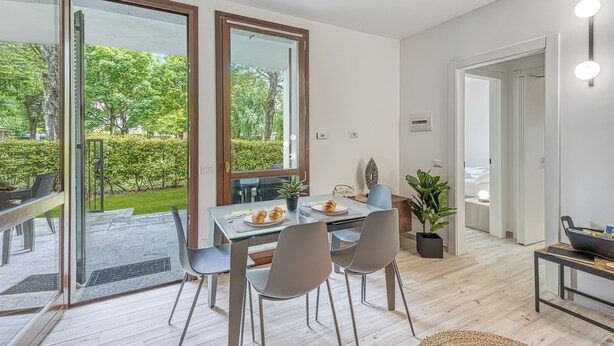 Un espace repas moderne avec une table en verre, des chaises grises et une vue sur un jardin grâce à de grandes fenêtres.
