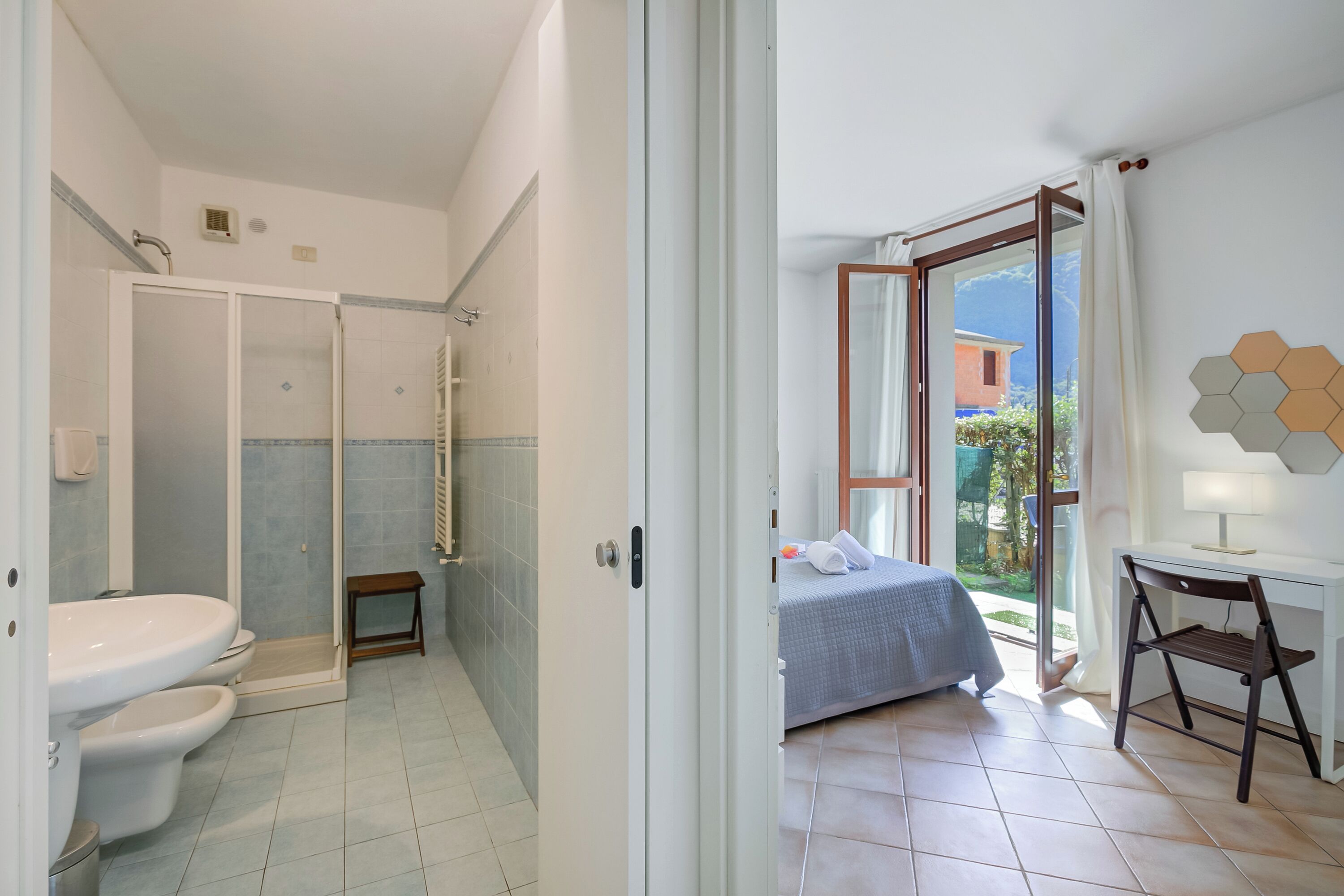 A bathroom with a shower, toilet, and sink. There is a bed with a blue blanket and a desk with a chair.
