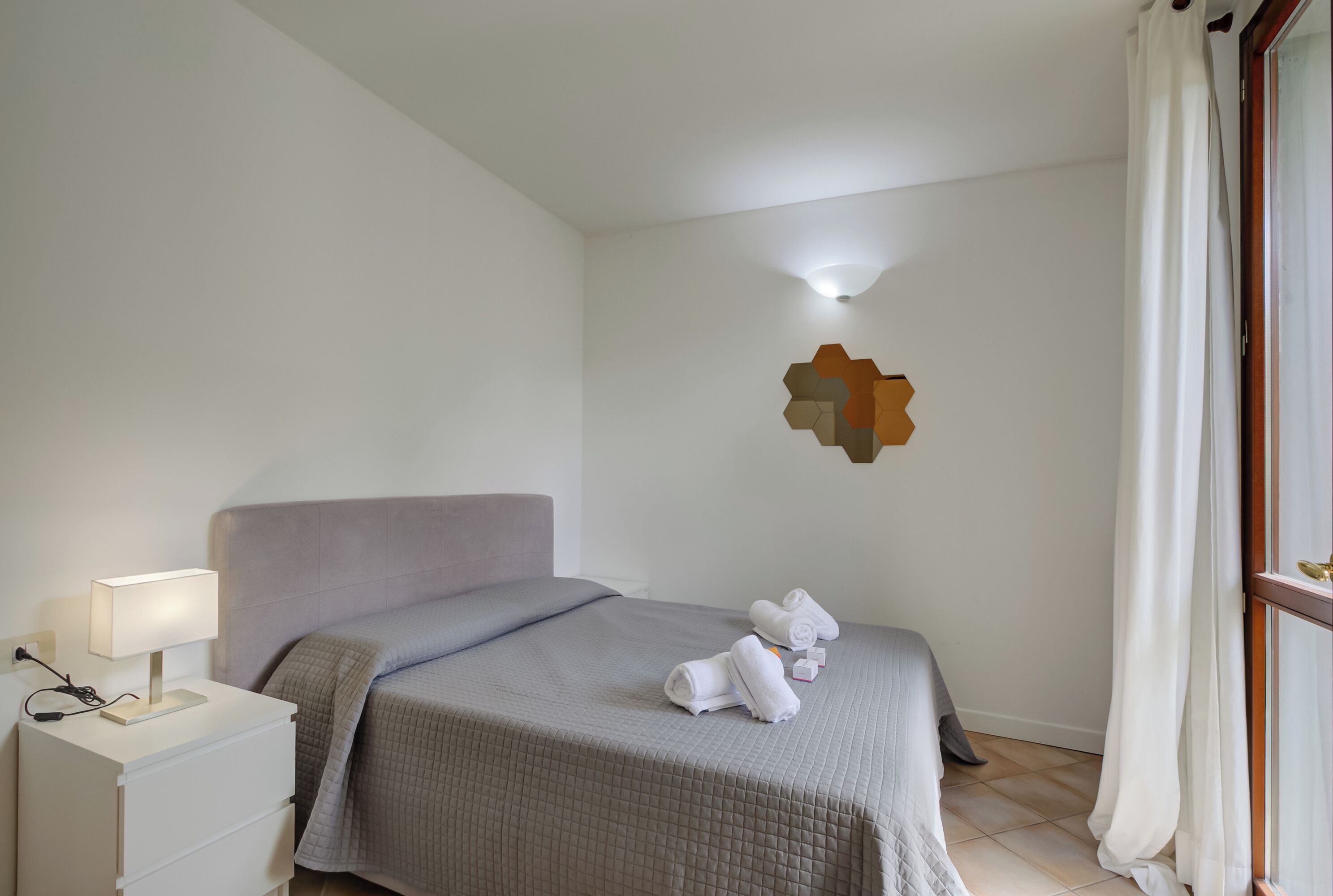 Ein ordentlich bezogenes Bett mit grauer Decke, ein Nachttisch mit Lampe und eine geometrische Wanddekoration.