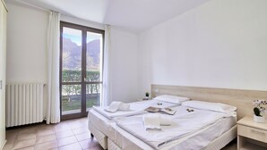 A hotel room with a large bed, a bedside table, a nightstand, a window with a view of greenery, and a radiator. - Porto Letizia Como 3, Porlezza, Italy (Porlezza)