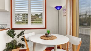 Een kamer met een ronde witte tafel, een potplant, een blauwe vloerlamp en een raam met witte luiken.