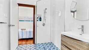Een moderne badkamer met een douche, toilet en wastafel. Een bed met een blauw-wit beddegoed is zichtbaar door een open deur.