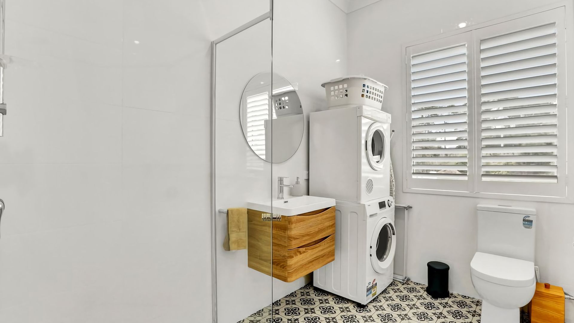 A modern bathroom with a glass door, a wooden vanity, a washing machine, a toilet, and a window with blinds.