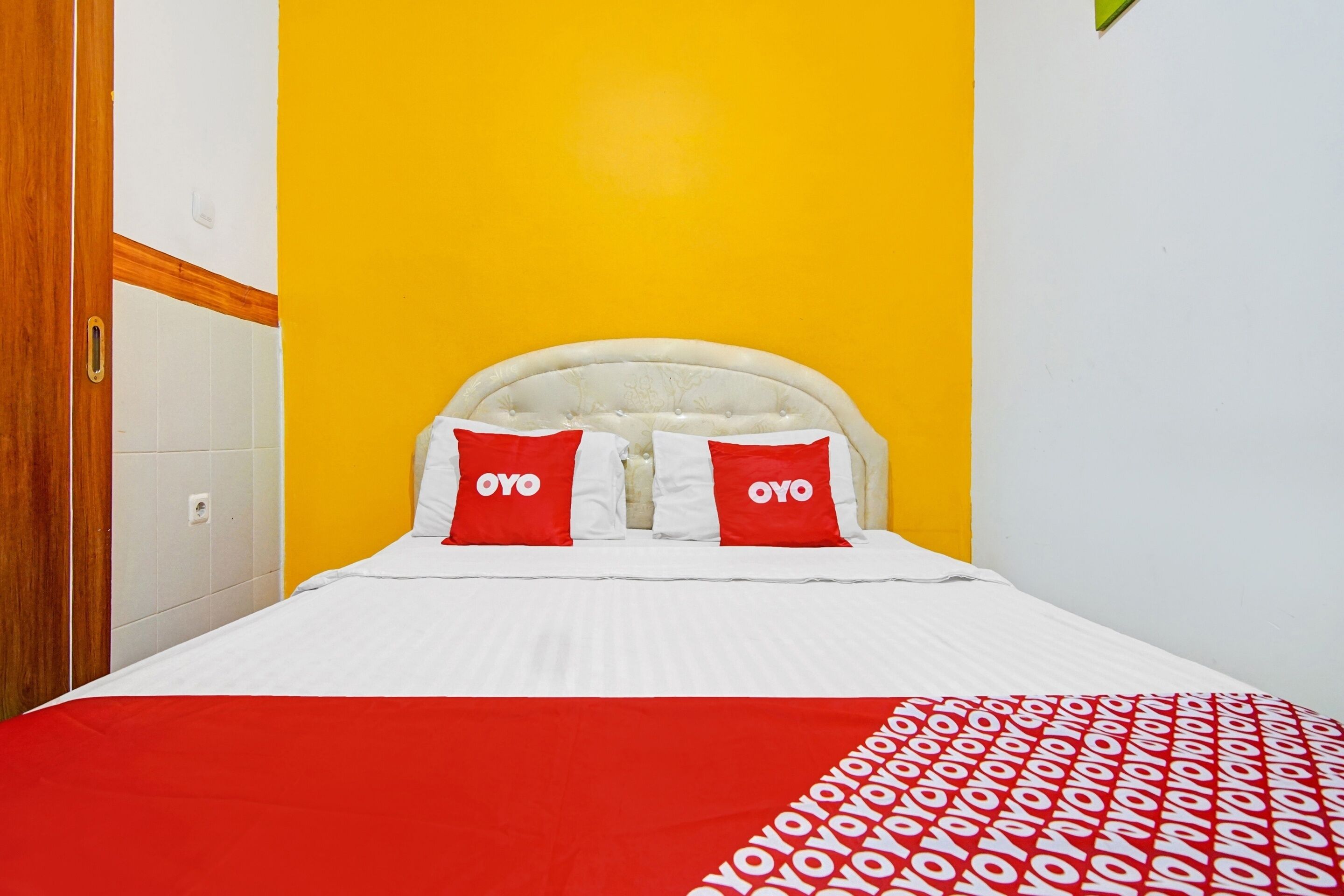 Un lit avec une tête de lit blanche, deux oreillers rouges ornés du texte « OYO » et une couverture rouge à motifs.