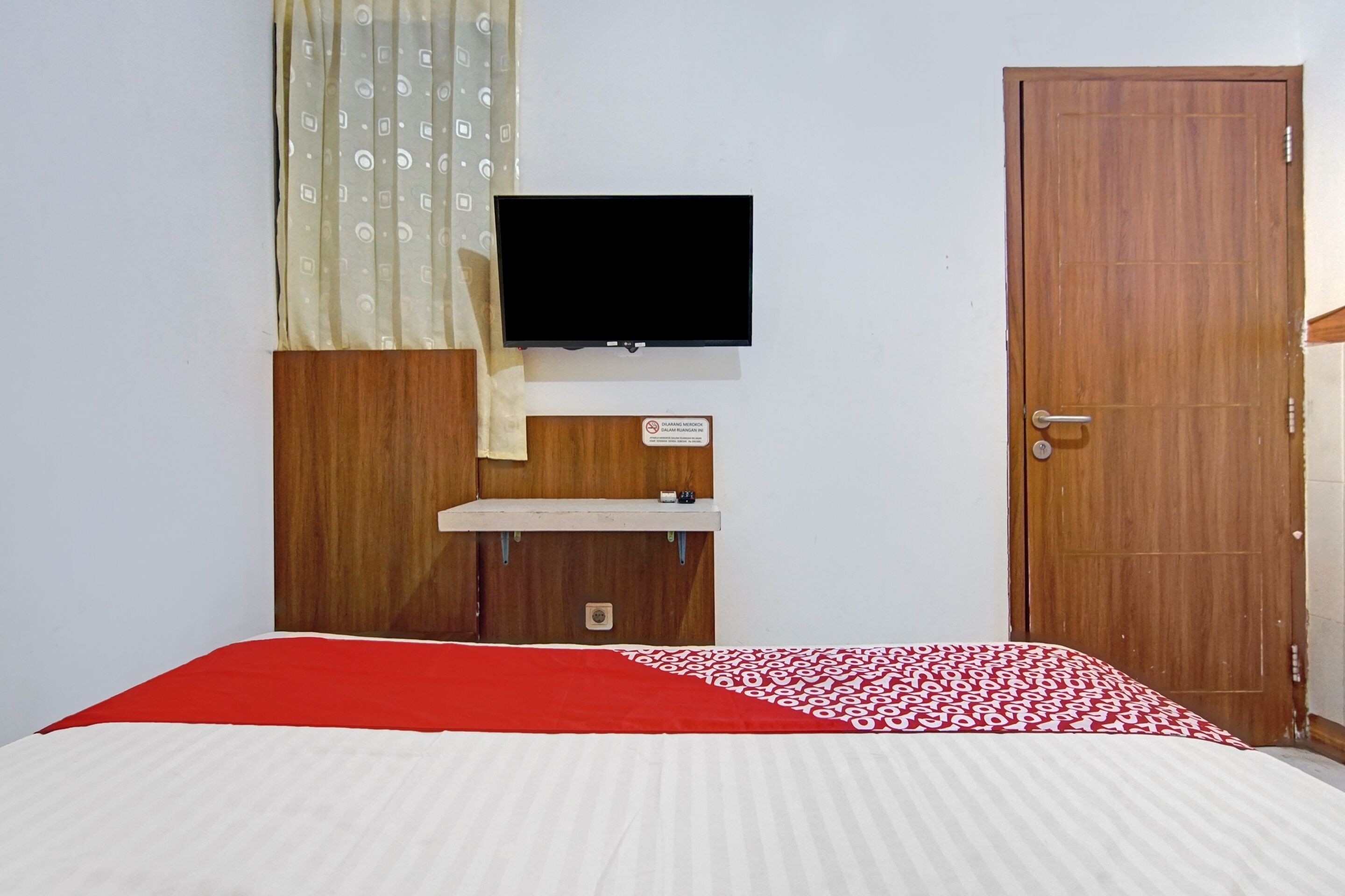 A bedroom with a bed, a television mounted on the wall, a wooden door, and a small wooden cabinet with a sign.