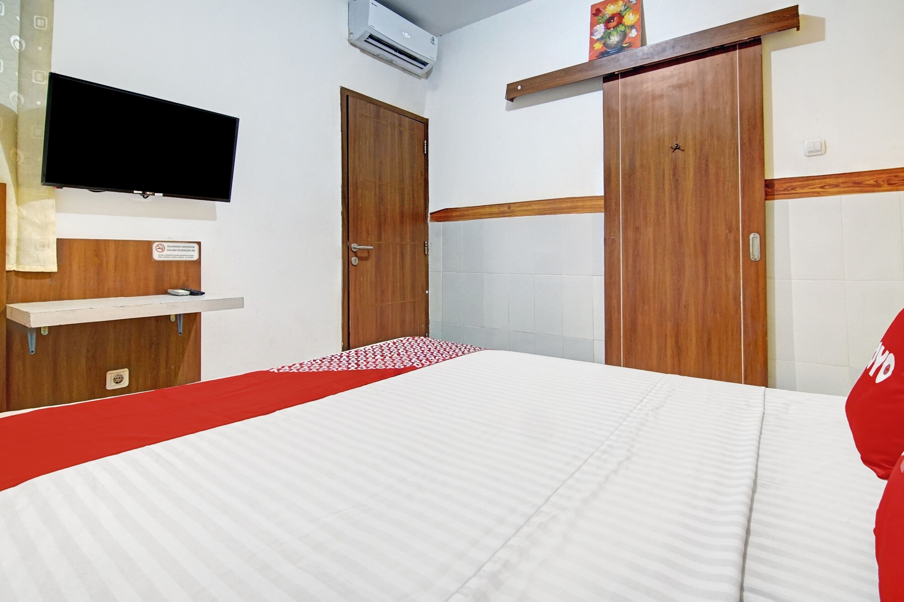 A hotel room with a bed, a TV mounted on the wall, a wooden wardrobe, and a red and white bedspread.