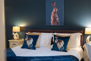 Quarto de hotel com uma cama grande, duas almofadas com ilustrações de coelhos, uma mesa de cabeceira com abajur e um quadro de coelho fixado na parede.
