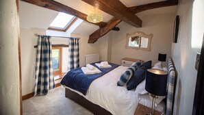 A bedroom with a sloped ceiling, a skylight, a bed with a blue blanket, a bedside table with a lamp, and a window with curtains. - Luxury cottage with enclosed garden and 2 dogs welcome (Castle Carrock)