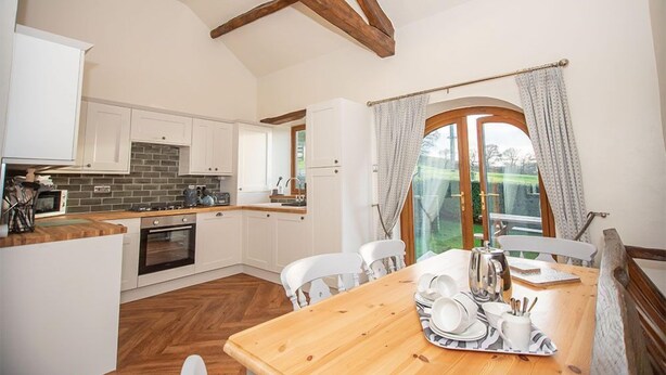 A modern kitchen with white cabinets, a brick backsplash, and wooden beams on the ceiling. There is a dining area with a wooden table set for two, and a large window with curtains. - Luxury cottage with enclosed garden and 2 dogs welcome (Castle Carrock)