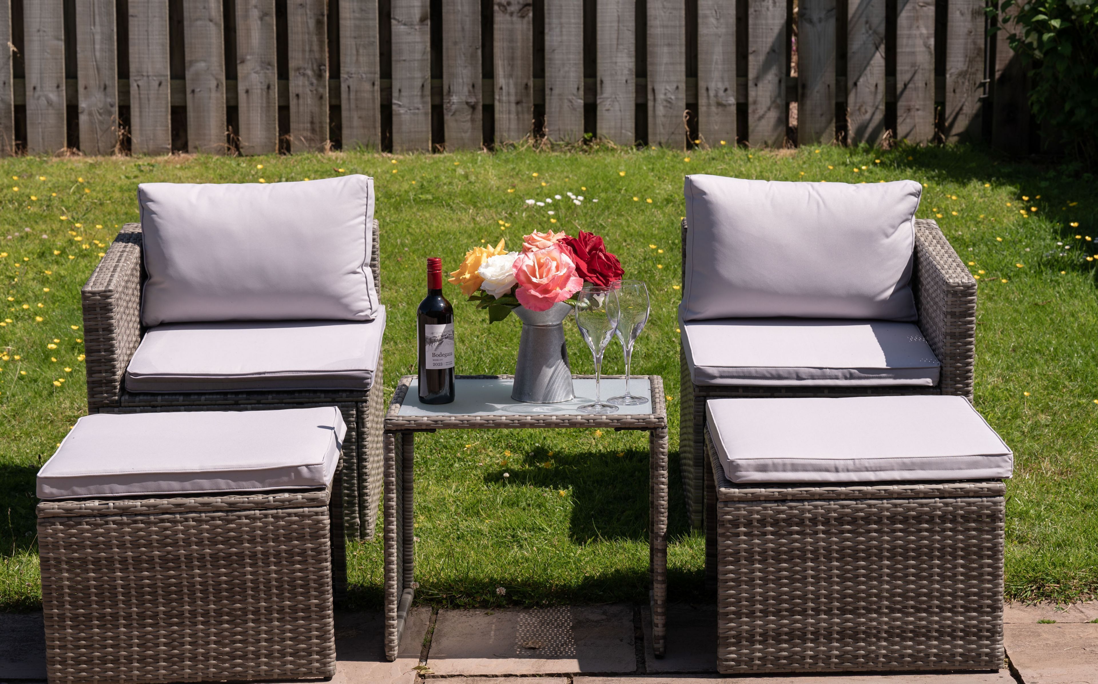 Two wicker chairs with cushions, a small table with a bottle of wine and a vase of flowers, set on a patio with a wooden fence in the background.