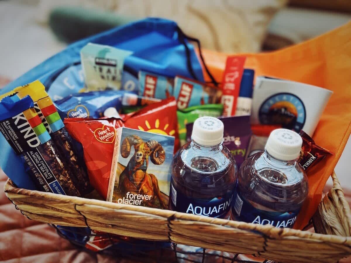Una cesta con varios artículos, incluyendo una tableta de chocolate, una caja de cereal, una botella de agua Aquafina y un libro titulado 'Forever Glacier'.