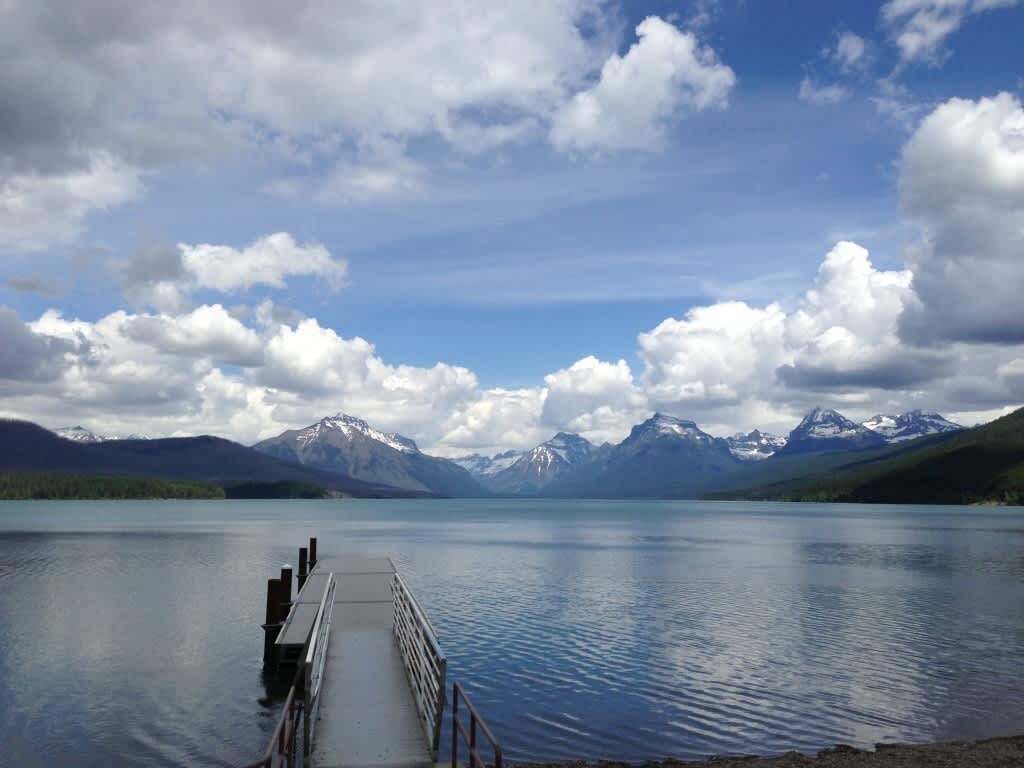 A calm lake with a dock extending into the water, surrounded by mountains and a partly cloudy sky.