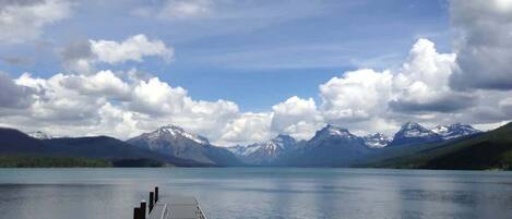 A calm lake with a dock extending into the water, surrounded by mountains and a partly cloudy sky.