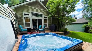 Un jacuzzi en un patio trasero con una casa, terraza y mobiliario exterior.