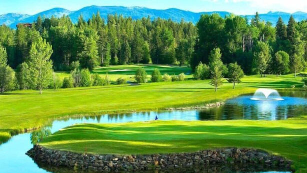 Un campo de golf con un estanque, árboles y montañas al fondo.