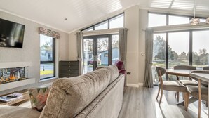 A modern living room with a sofa, a flat-screen TV, a fireplace, and large windows offering a view of the outside. - 3 bedroom accommodation in Faringdon (Faringdon)