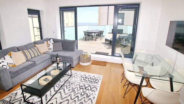 A modern living room with a grey sofa, a glass coffee table, and a dining area with a glass table and chairs. - Worthing Beach, Sea View, Balcony | 2 Apartments (West Sussex)