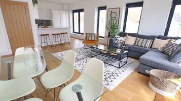 Een moderne woonkamer met een bank, salontafel en stoelen. Achtergrond is een keuken met witte kasten en een eethoek met een glazen tafel en stoelen.
