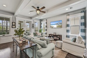 Un salon spacieux avec un canapé d’angle, une table basse et un téléviseur. De grandes fenêtres sont agrémentées de rideaux, et la pièce est équipée d’un ventilateur de plafond et d’une cheminée.