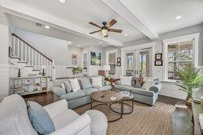 A spacious living room with a high ceiling, a ceiling fan, and a staircase leading to another level. There are two sofas, a coffee table, and a bookshelf.