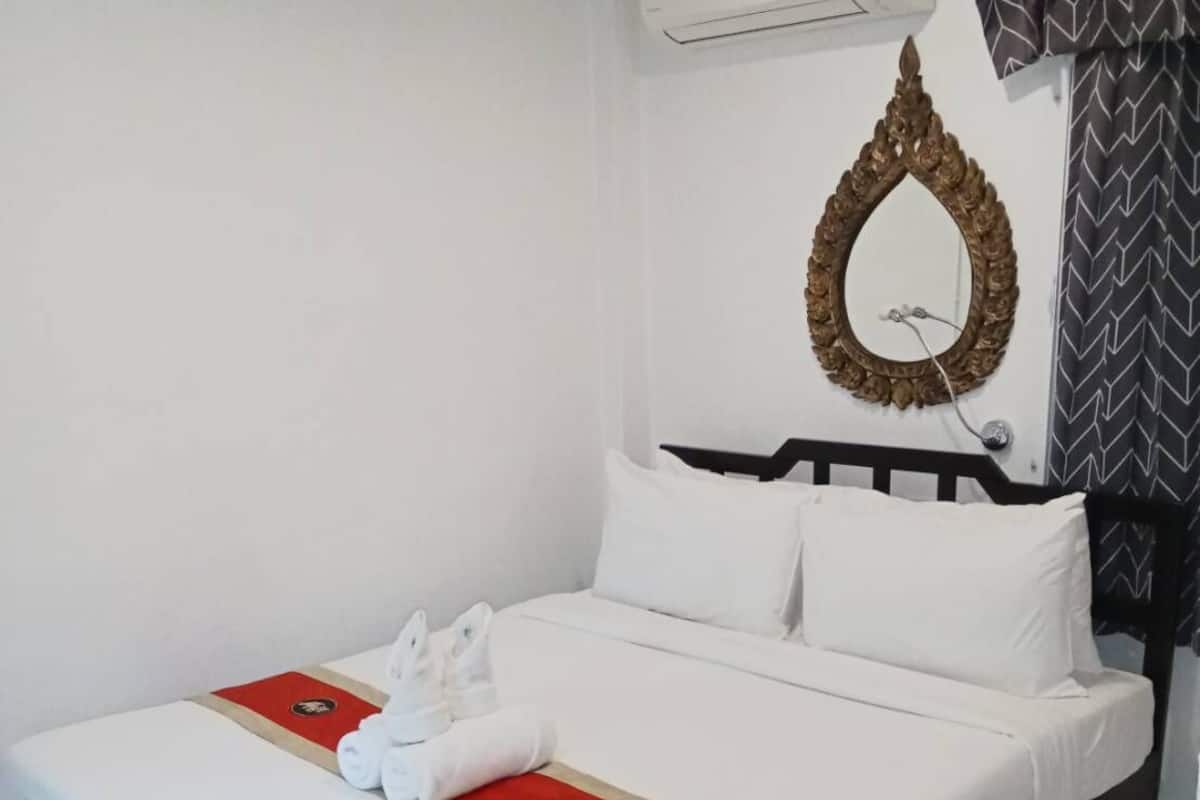 A hotel room with a bed, white bedding, a red and gold bed runner, a wall-mounted air conditioner, a decorative mirror, and a patterned curtain.