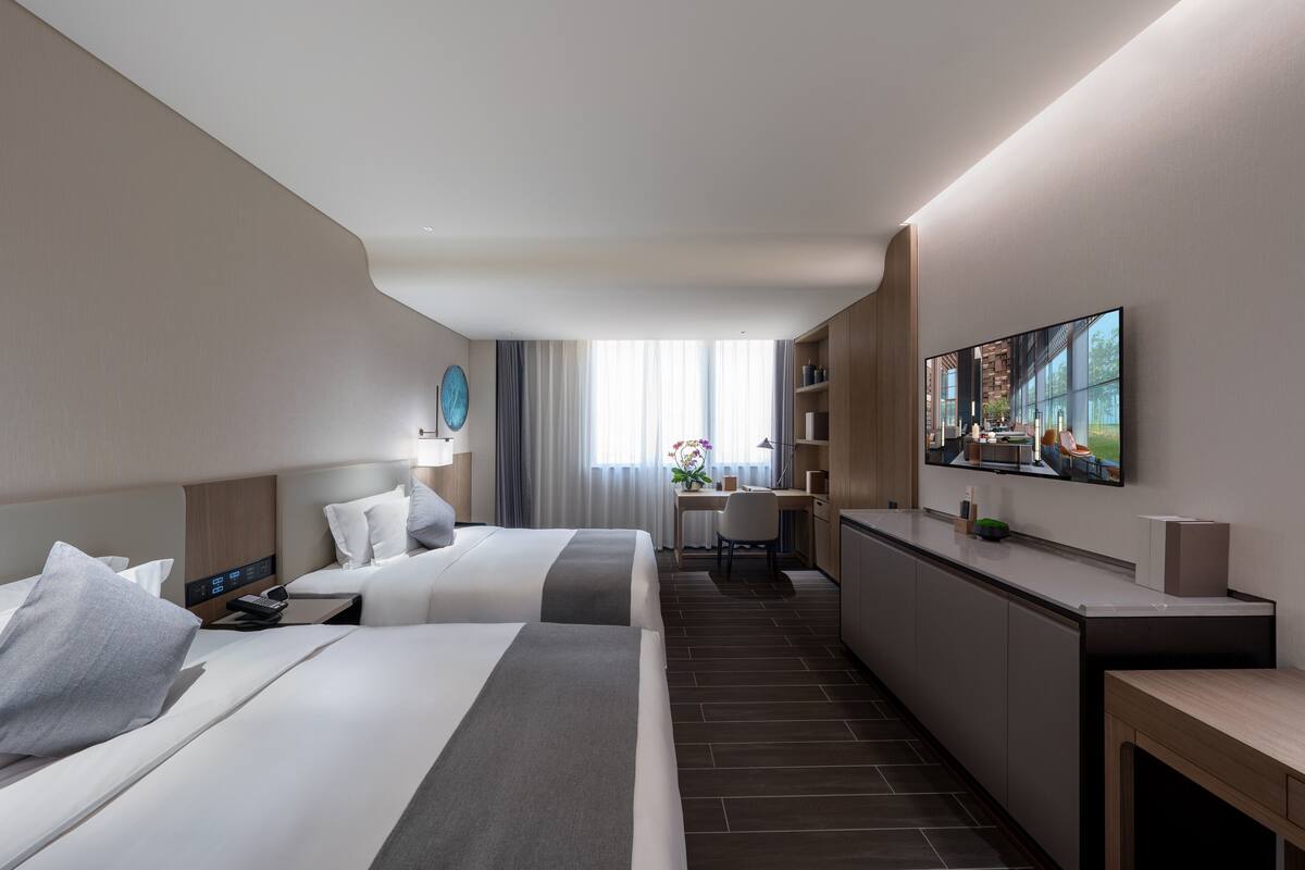 Une chambre d’hôtel moderne équipée de deux lits, d’un bureau, d’une télévision à écran plat et offrant une vue sur la ville grâce à de grandes fenêtres.