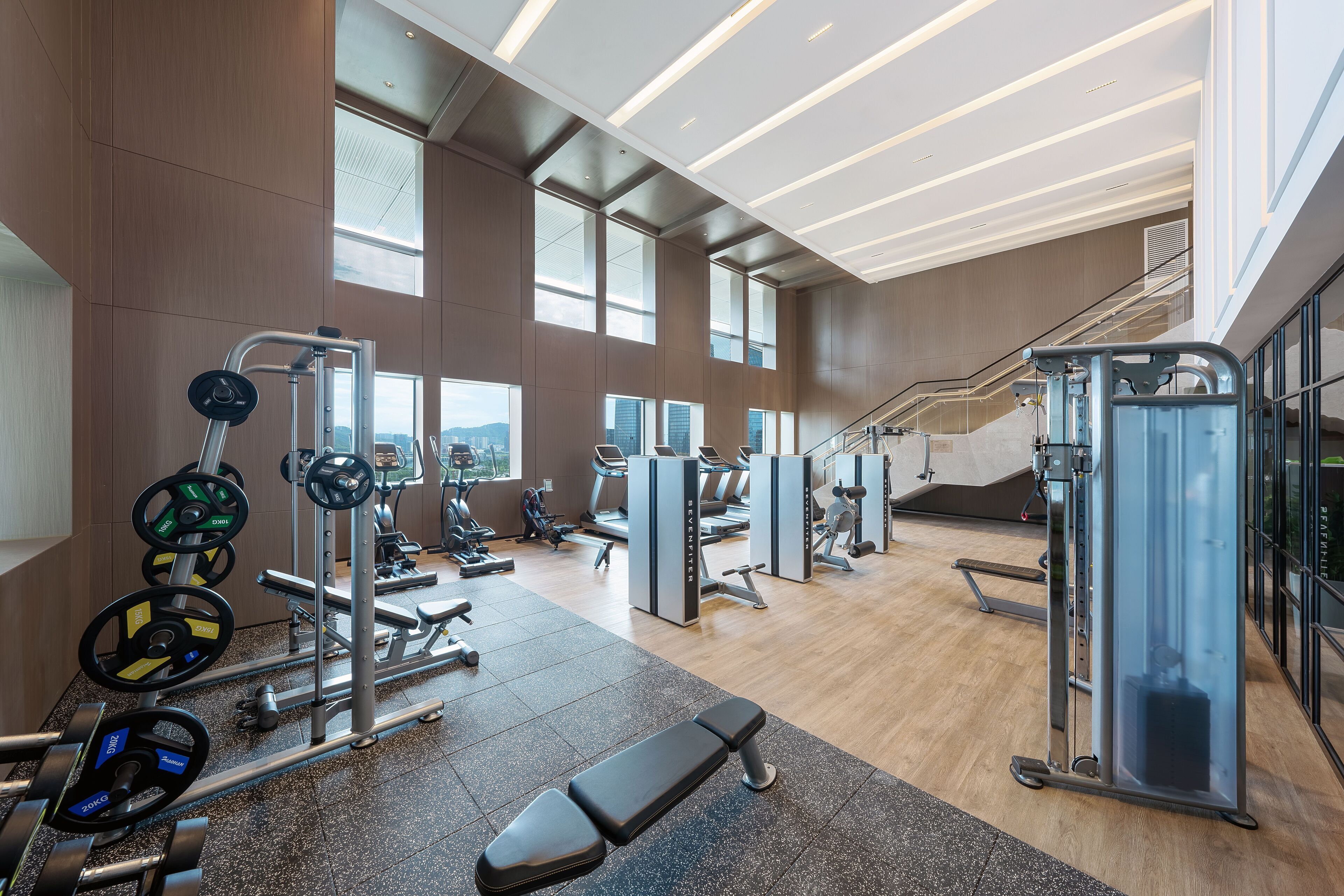 A modern gym with various exercise equipment, large windows, and a staircase.