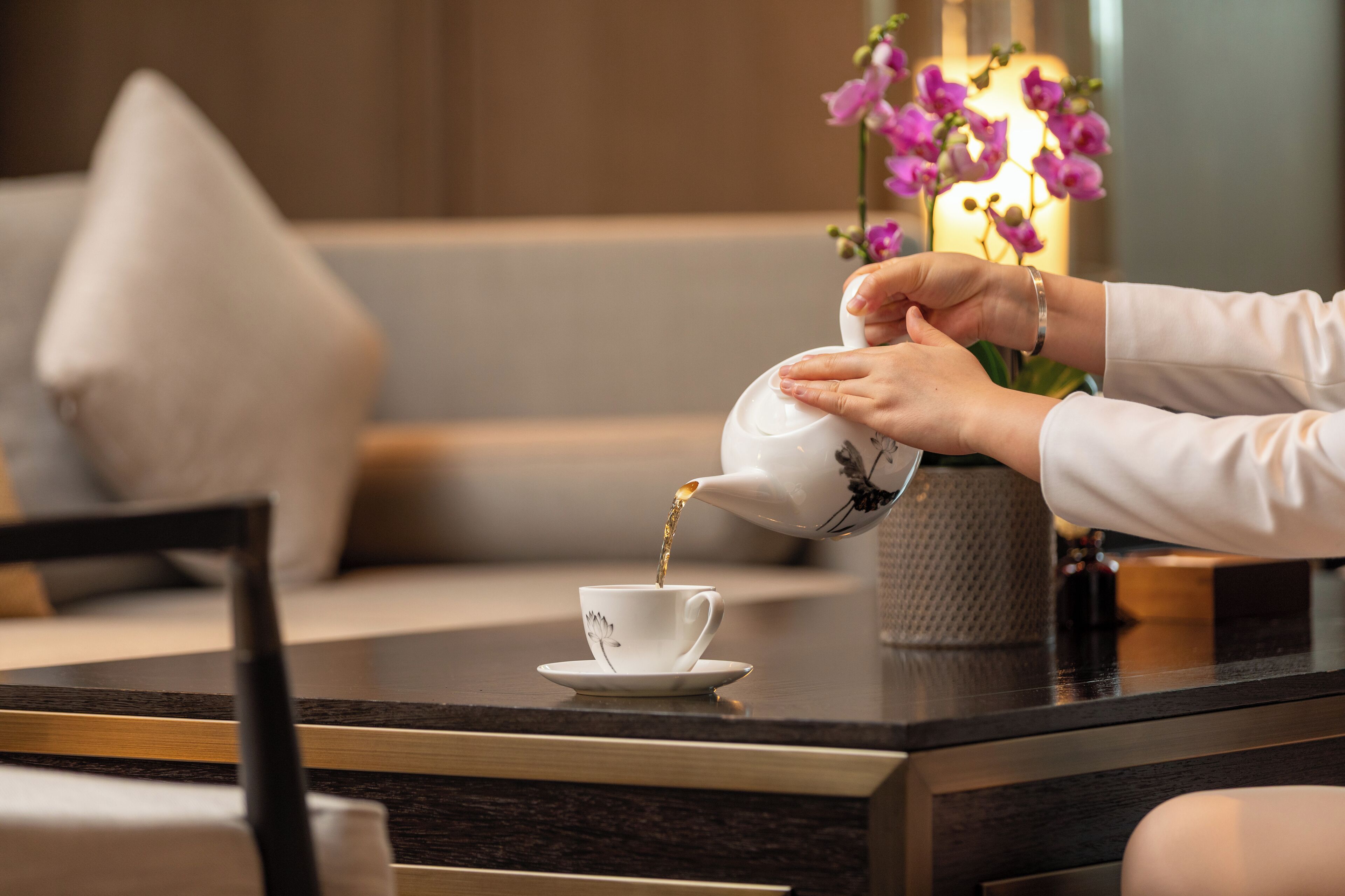 Una persona sirve té en una taza sobre una mesa, con un florero en el fondo.