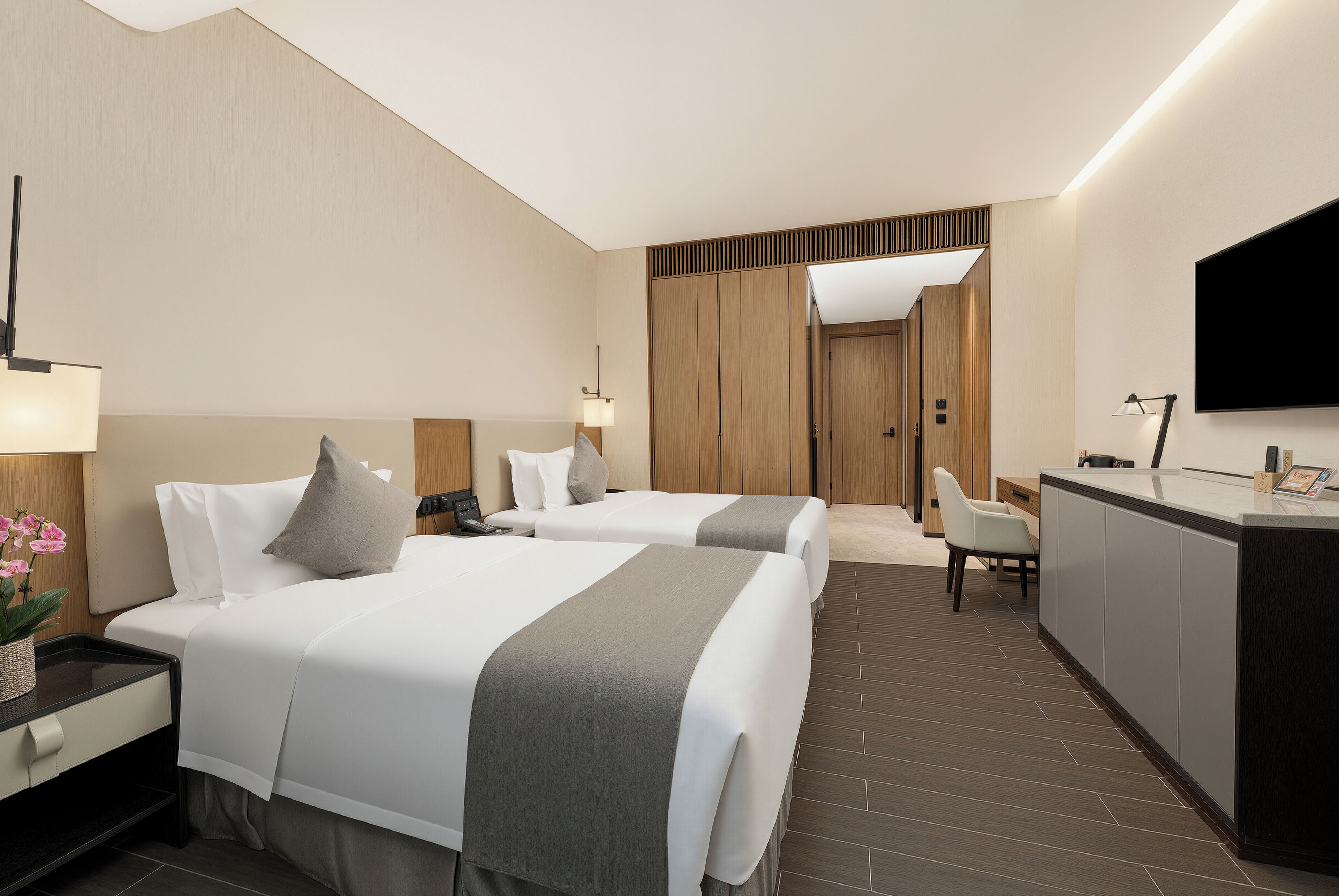A hotel room with two beds, a desk, a chair, a TV, and a reception area.