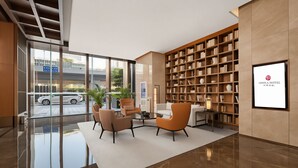Eine moderne Hotelhalle mit Glastür, einer Sitzecke mit Sesseln, einem Bücherregal und einem gerahmten Logo an der Wand.