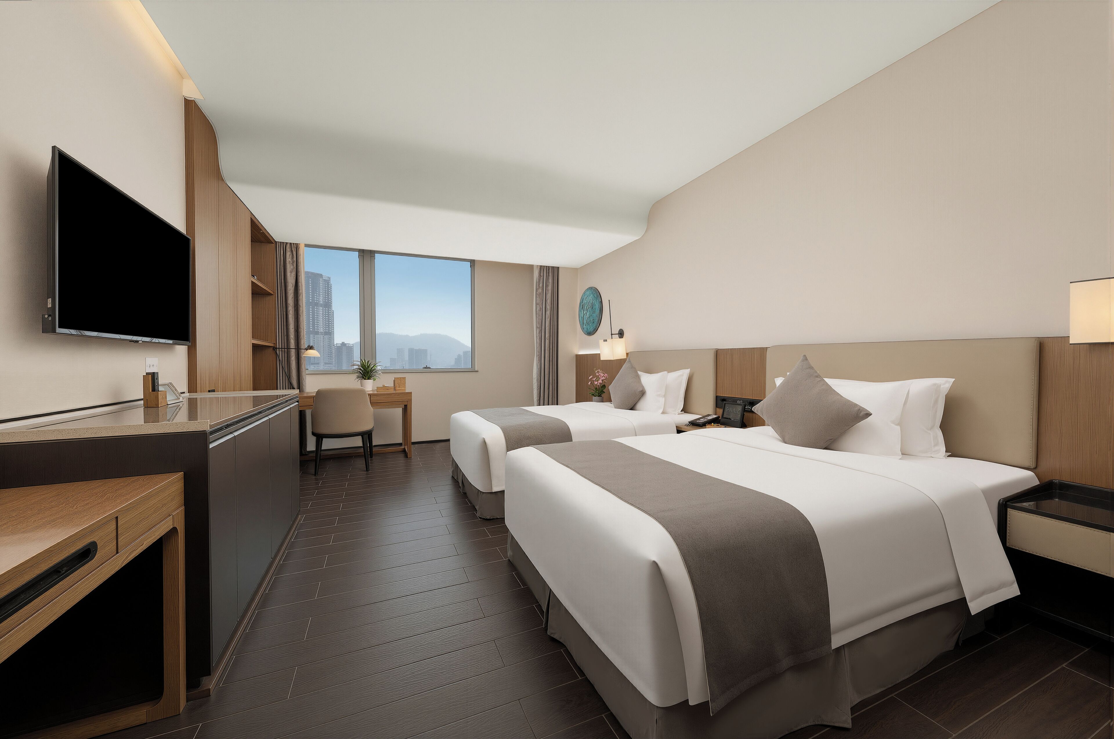 A hotel room with two beds, a desk, a TV, and a view of the city.