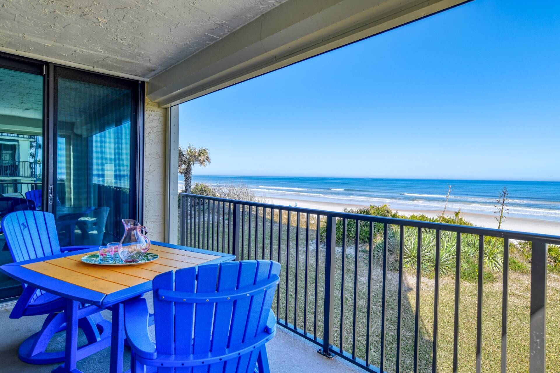 Ein Balkon mit einem Holztisch und blauen Stühlen, der Blick auf einen Strand mit klarem, blauem Wasser und sandigem Ufer gewährt.