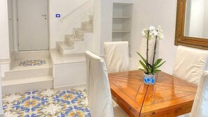 Un coin repas avec une table en bois, des chaises blanches, un vase avec des fleurs blanches, un escalier avec des rampes blanches et un sol carrelé aux motifs bleus et jaunes.