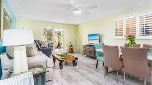 A living room with a ceiling fan, a TV, a dining area with chairs, and a view of the outdoors through a sliding door. - Lulu's Cozy Beach Cottage #1 | 5 Min. Walk to Beach | Dog Friendly | Pool | $185 Free Activities! (Sarasota)