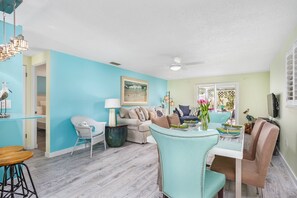 A spacious living room with a light blue wall, a ceiling fan, a dining area with a table set for two, and a large window providing natural light. - Lulu's Cozy Beach Cottage #1 | 5 Min. Walk to Beach | Dog Friendly | Pool | $185 Free Activities! (Sarasota)