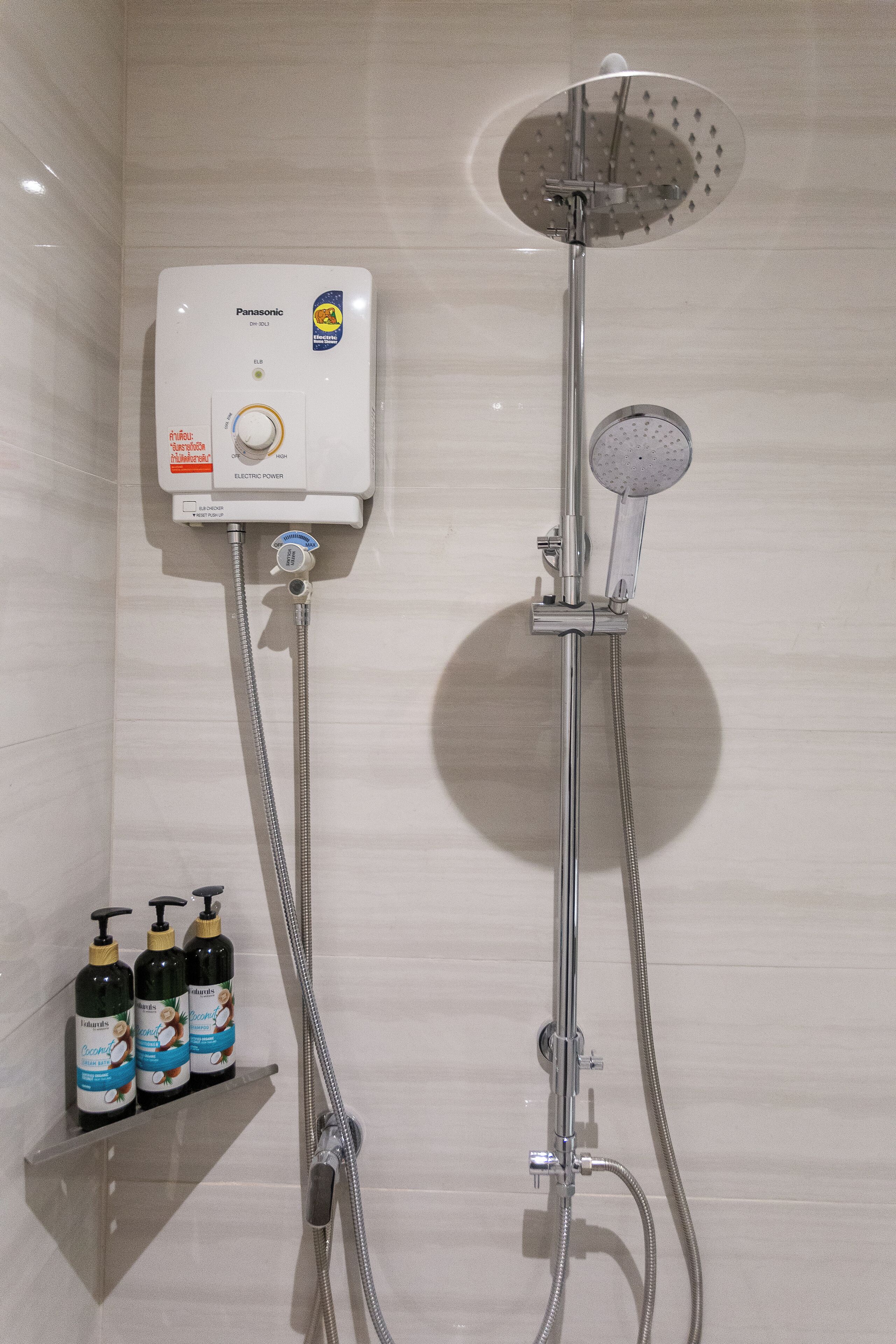 Badeværelse med en Panasonic elbruser, et håndholdt brusehoved og en hylde med tre shampoo flasker.