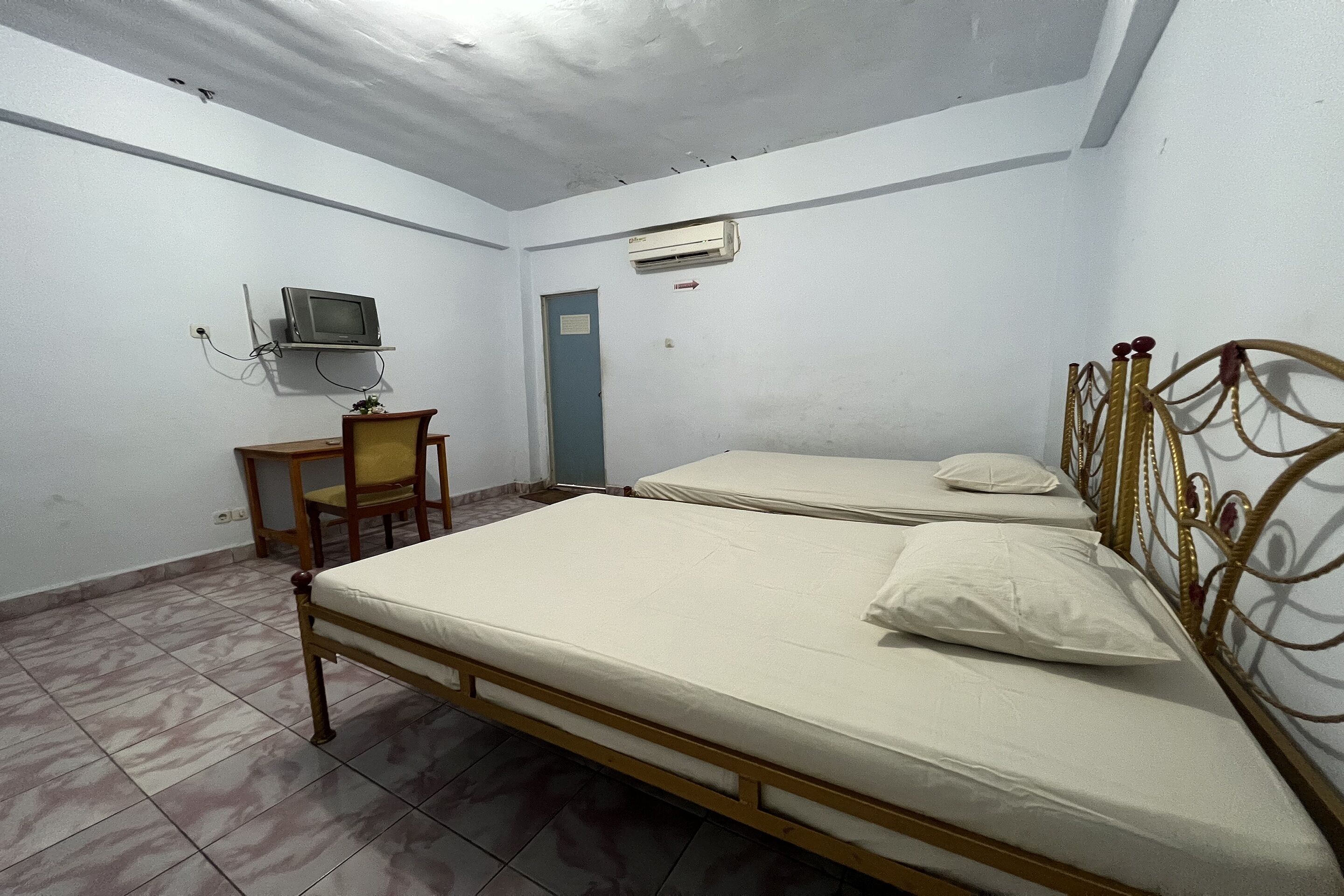 A room with two beds, a desk with a chair, a television mounted on the wall, and an air conditioning unit.
