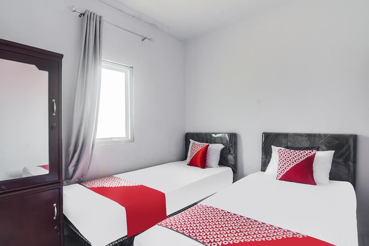 A hotel room with two beds, a wooden wardrobe, a window with curtains, and a plain white wall.