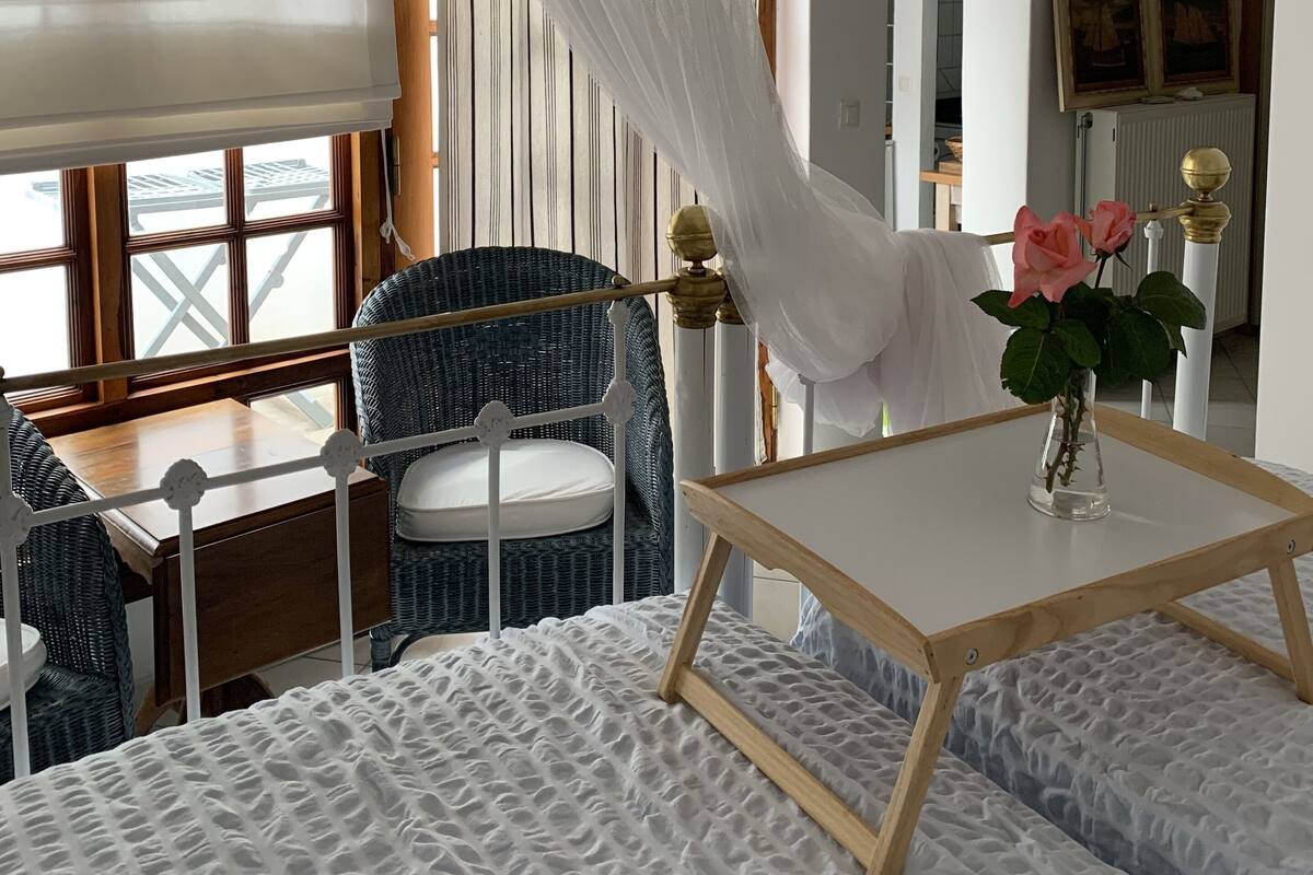 A bedroom with a large window, a bed with a textured white cover, a bedside table with a vase of flowers, and a chair with a white cushion.