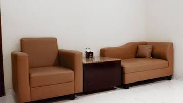 Una habitación con un sofá de cuero marrón y un sillón a juego, una pequeña mesita de noche oscura con una botella y una caja de pañuelos, y un piso con baldosas blancas.